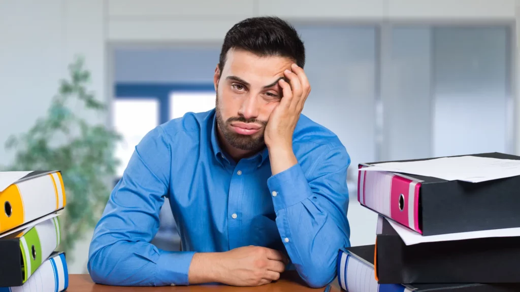 Un homme fatigué sur son bureau et entouré de classeurs qui s'empilent.